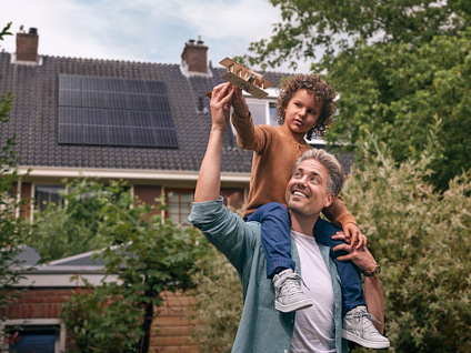 Man met kind op schouders die beide een speelgoedvliegtuig vasthouden voor een huis met zonnepanelen