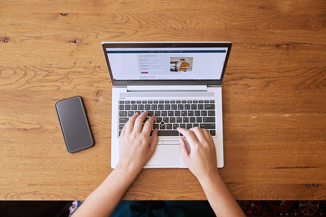 Handen die aan het typen zijn op een opengeklapte laptop met daarnaast een telefoon op tafel