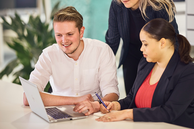 Drie collega's die samen werken op een laptop. Twee zitten er en eentje kijkt over hun schouders mee.