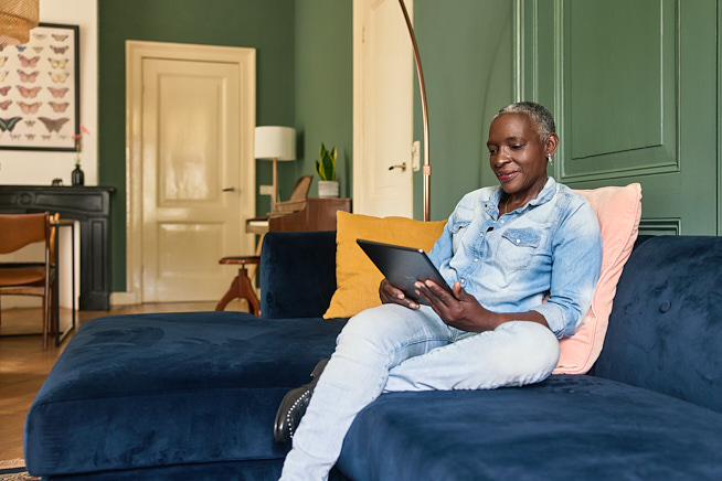 Lachende vrouw die zit op de bank in haar woonkamer met een tablet in haar handen